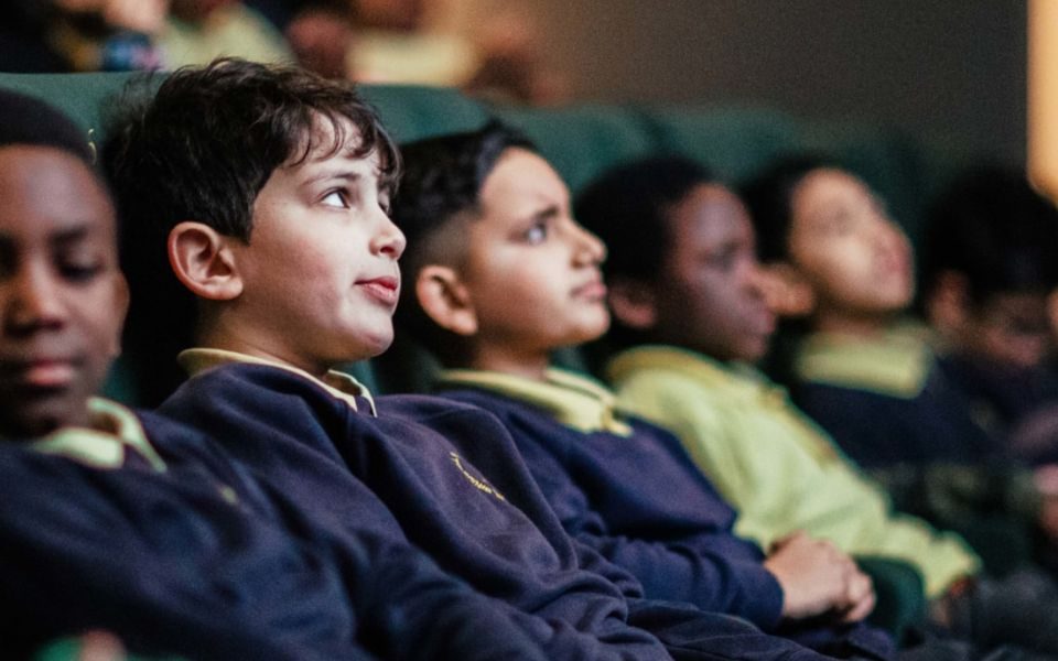 A row of primary schoolchildren in blue and yellow uniforms sitting in green cinema chairs watching something on the big screen