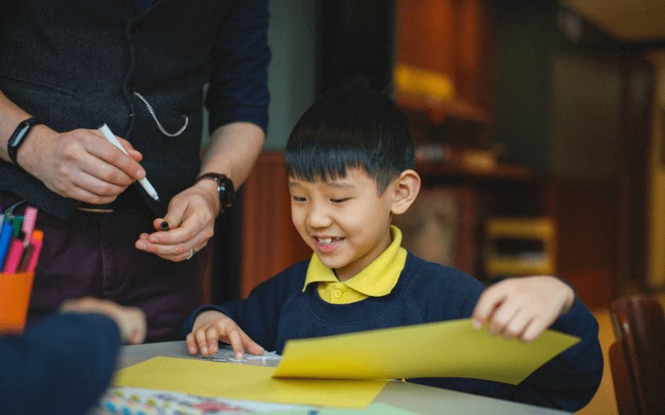A primary schoolchild sits smiling at a desk with a piece of yellow paper