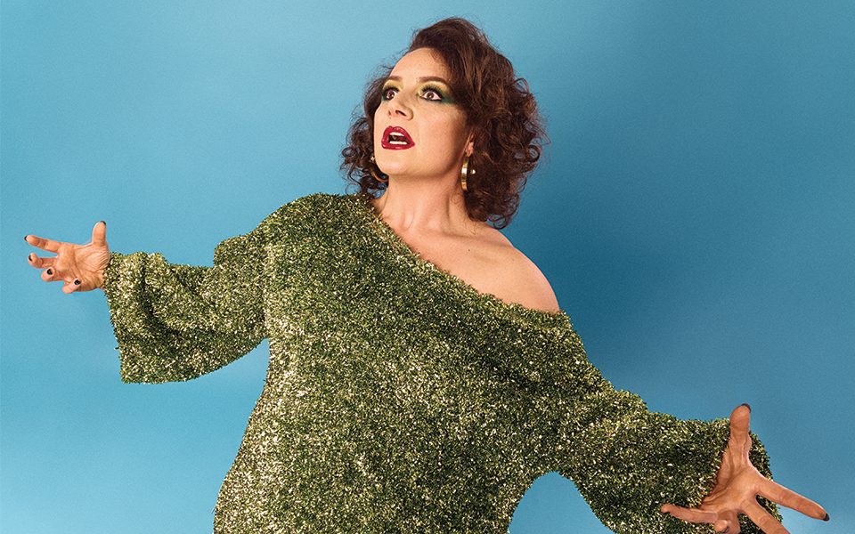 Jessica Fostekew pulls a shocked expression with outstretched arms, leaning back slightly. She wears strong make-up, hoop earrings and a glittery top.