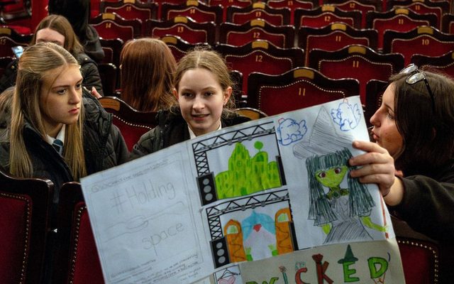 Three students sit in Stalls at The Varieties waiting for their turn to present and holding up a show design poster