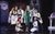 Rocky Horror Show Frank-N-Furter in a laboratory wearing a green robe, fishnets and heels points ahead and is surrounded by the Rocky Horror Show cast wearing white lab coats and aprons looking shocked.