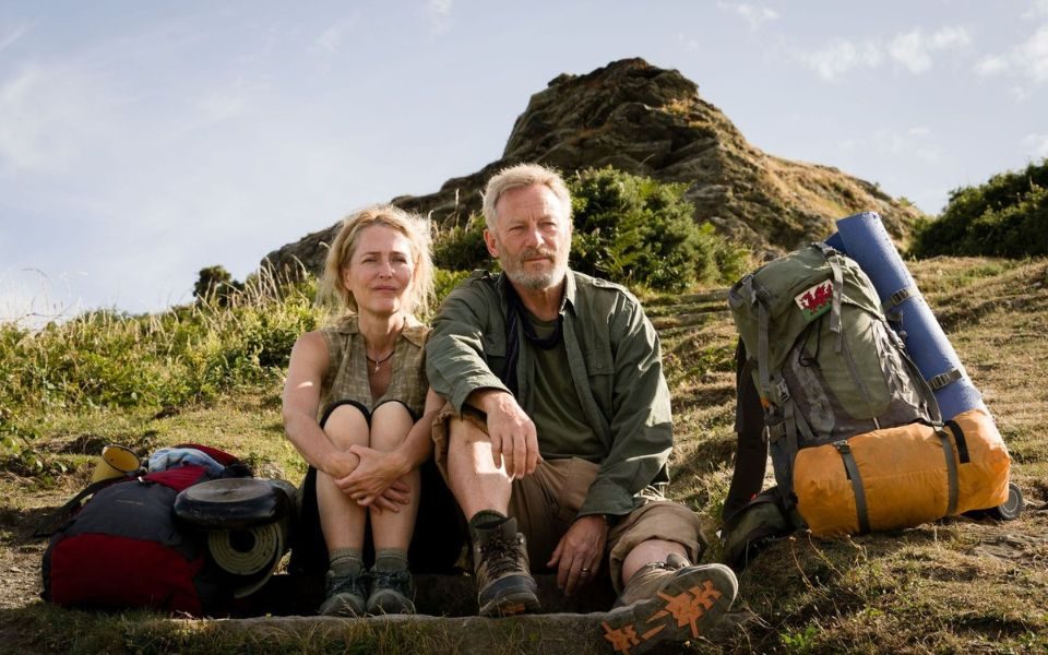 Gillian Anderson and Jason Isaacs sat on a grassy dune in summery hiking gear with a trekking rucksack next to each of them.