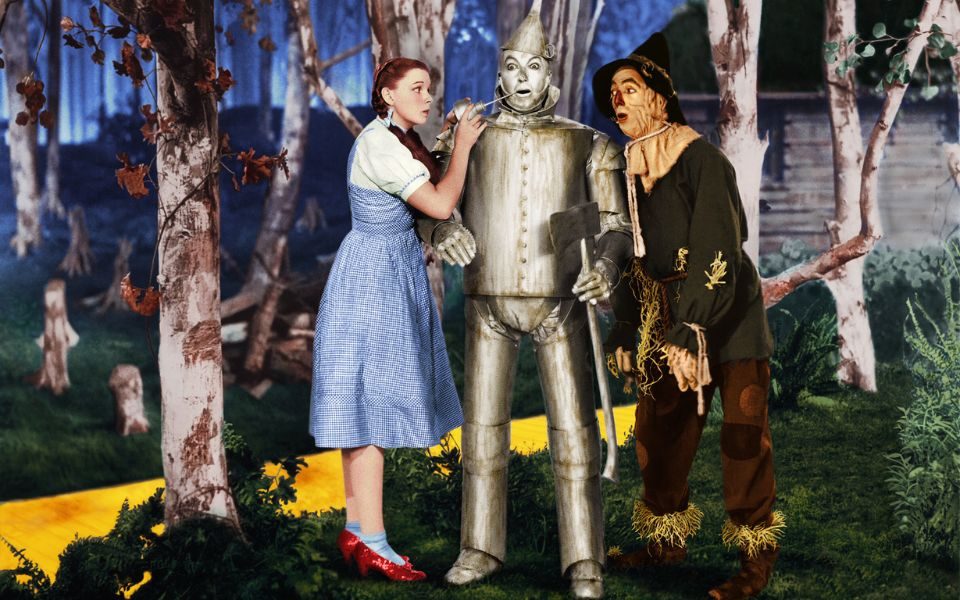 The silver tin man with Dorothy in her blue dress and red heels to his left and the scarecrow to his right. The yellow brick road can and trees are in the background.
