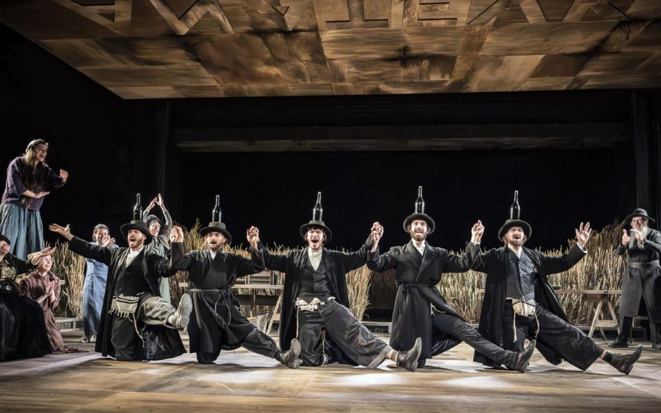 Ensemble perform a traditional wedding dance, with bottles balanced atop their hates, as they strike poses with one knee to the ground