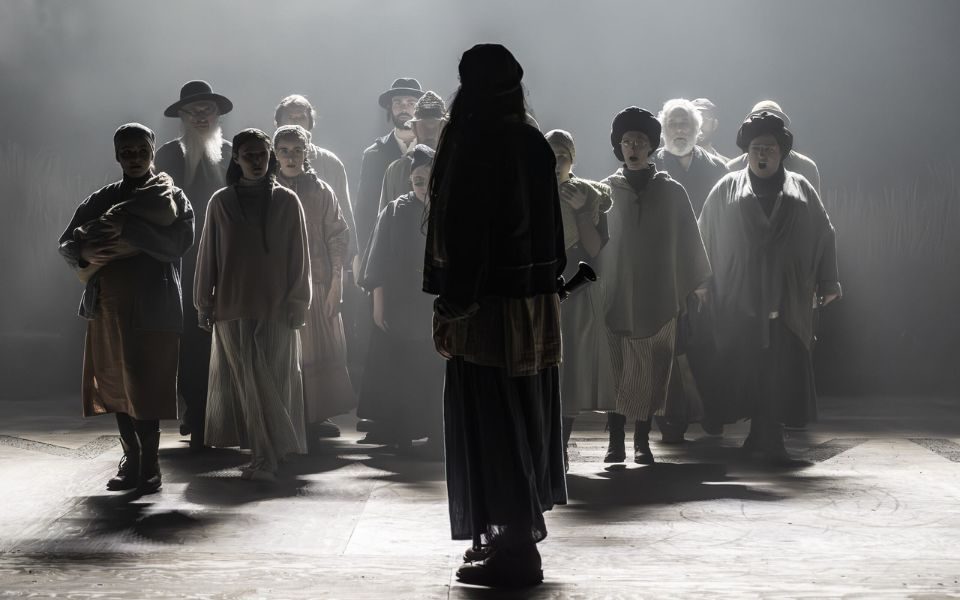 Ensemble walking forward on a dark stage as a silhouetted figure looks back at them