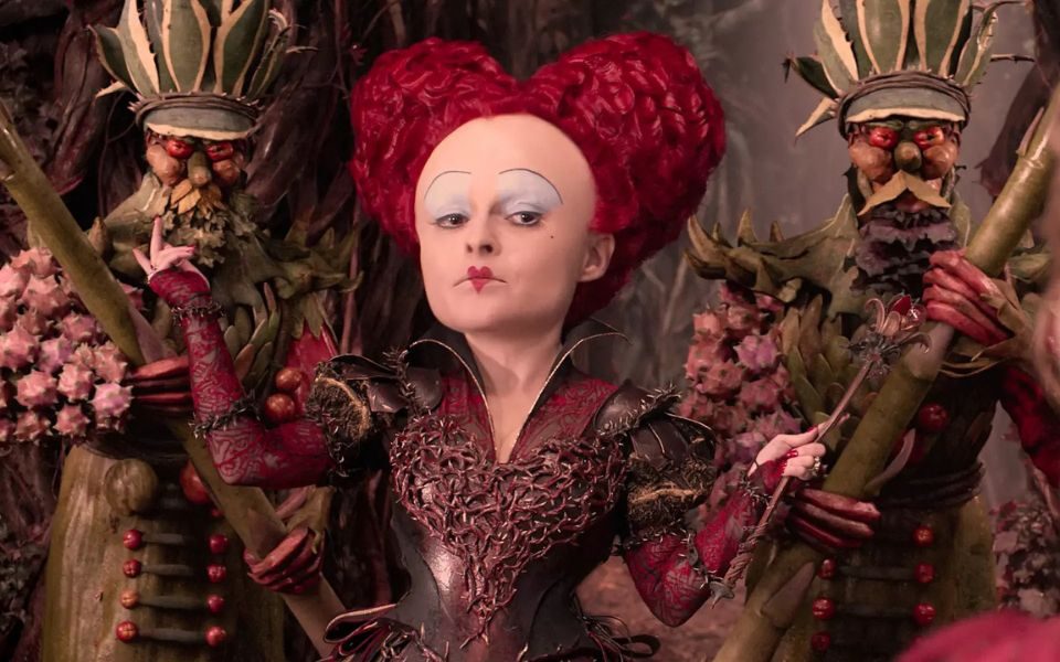 The red-haired Queen of Hearts wears an extravagant but spiky metallic corset and gestures at two guards accompanying her, both seemingly made of plants and holding large staffs.