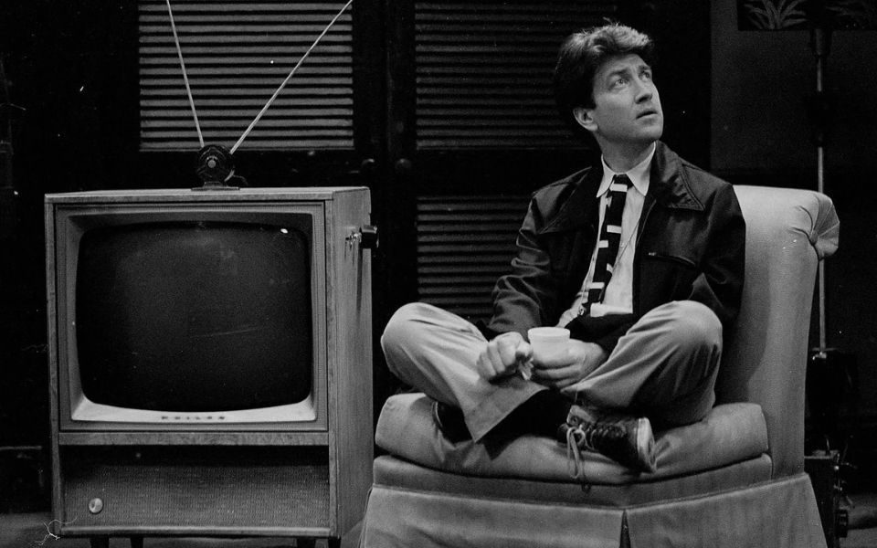 Black and white still. David Lynch sits suited and cross-legged atop a cushioned chair beside an old-school television set.