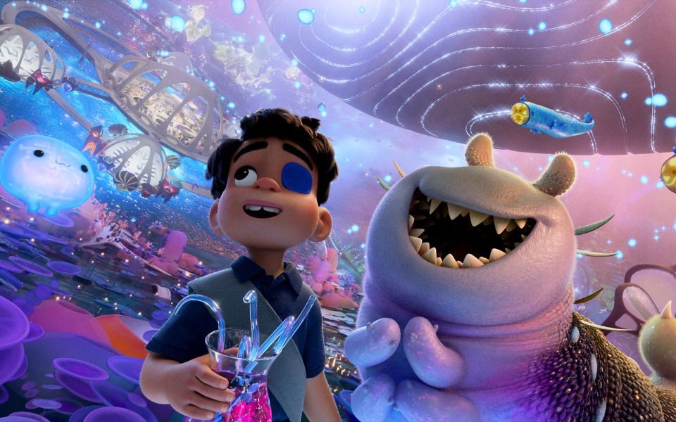 Animated digital still. A young boy wearing an eye patch stares up in wonder at a galaxy full of spiralling space stations, glowing orbs and smiling jellyfish-like creatures. He stands beside a friendly but sharp-toothed slug-like alien.
