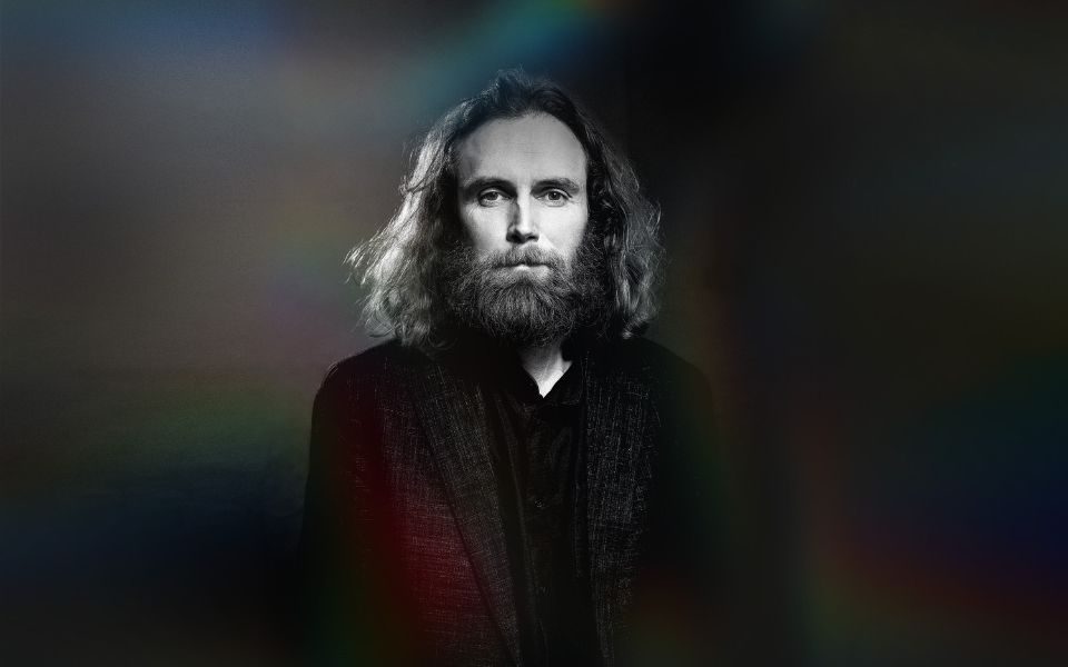 In black and white, Rob Auton with dark shoulder-length hair and beard dressed in a black suit stands alone and serious against an empty backdrop.