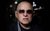 Shaun Ryder Shaun Ryder holds a neutral gaze, donning a pair of Ray Bans and navy jacket. The background is blurred but busy with lights and activity.