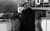 The Elephant Man In black and white, a cloaked man with a sack masking his face walks across a boat, surrounded by staring bystanders in Victorian-era clothing and top hats.