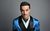 Troy Hawke Moustachioed Troy Hawke wears a shiny blue floral smoking jacket and matching neck tie against a plain grey background.