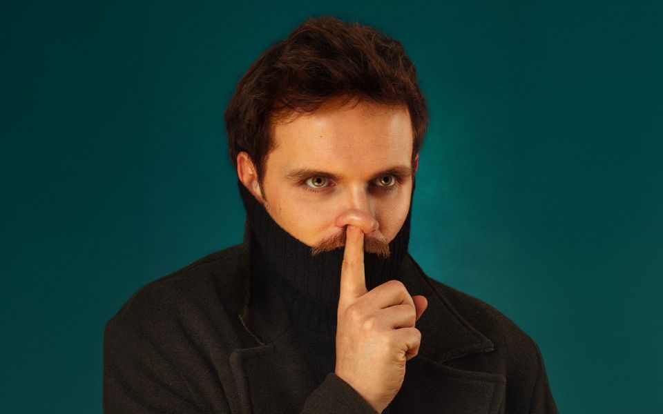 Vittorio Angelone with a turtle neck pulled up his mouth and his finger over where is mouth would be in a 'be quiet' gesture.