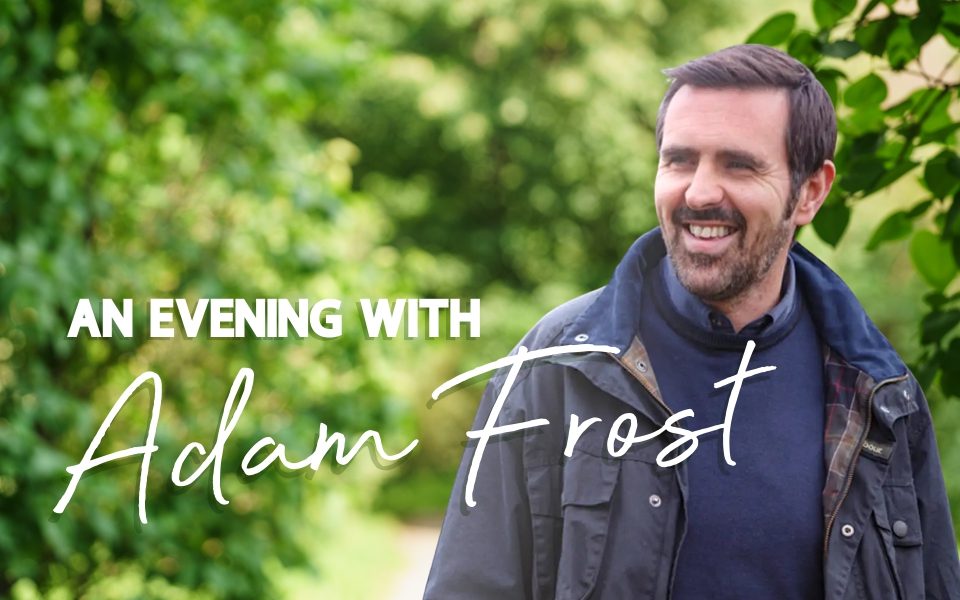 A smiling Adam Frost in navy knitwear and a coat casually looks off-camera in a luscious green garden. Text reads: An Evening with Adam Frost.