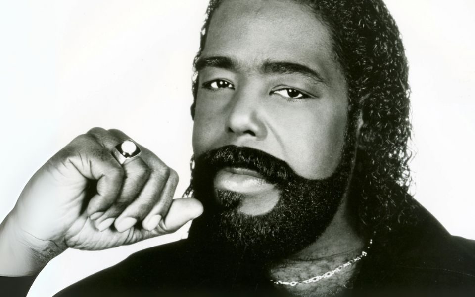 Barry White photographed in black and white, set against a blank backdrop. He has a tight perm and carefully shaped beard, wearing a ring and chain necklace.