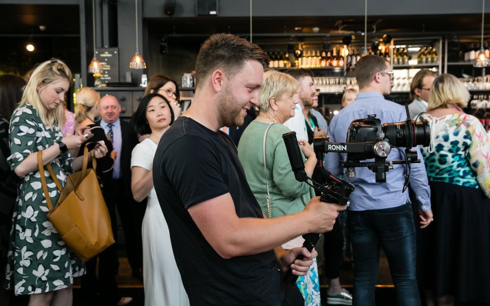 In profile, Chris Coote operates a camera on a gimble in a busy bar setting.