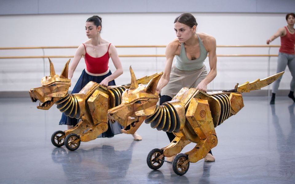 Two puppets on wheels are being operated in sync by two puppeteer dancers in a rehearsal studio.