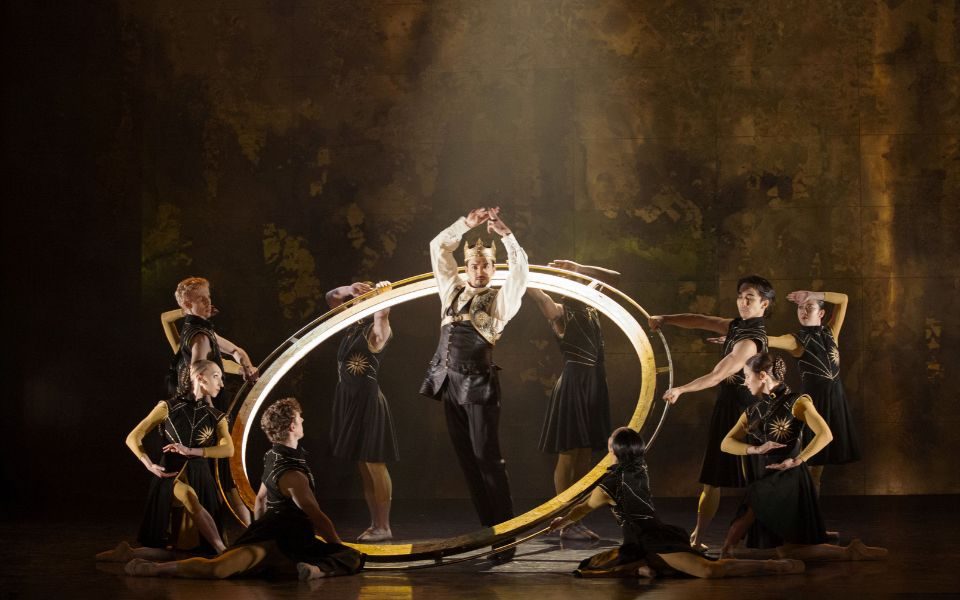 A king stands within a large golden halo handled by multiple ballet dancers.