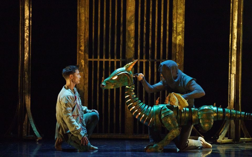 A performer knelt on stage, facing a glowing dragon puppet operated by a cloaked figure.