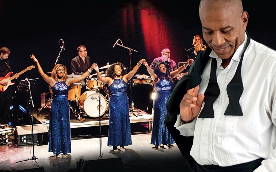 Roy Hemmings stands in the foreground in a relaxed pose, a loose bowtie round his collar and suit jacket flung on his shoulder. Three singers in sequined blue dresses stand proudly in front of a full band on-stage.