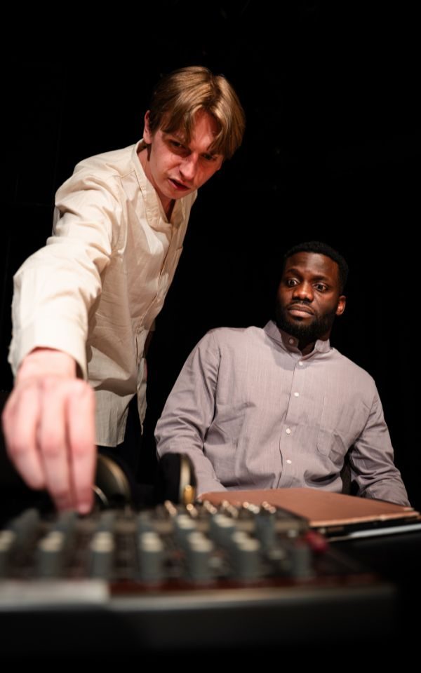 One man leans towards a mixing desk, whilst the other sits back and observes.