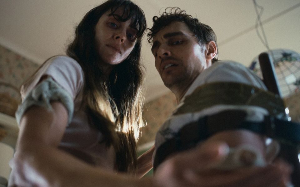 Shot from a low angle, sweaty and stressed couple Alison Brie and Dave Franco look down at something out of view in Alison Brie's hand. A tourniquet bandage is wrapped round her arm. He is taped to a chair.