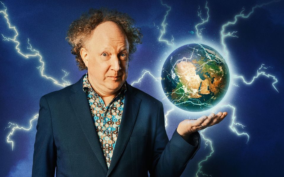 Andy Zaltzman stands in a suit jacket and patterned shirt. A globe emitting electricity and lightning bolts hovers above his outstretched hand.