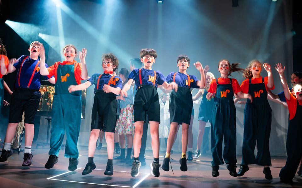 A line of children aged 8-11 are lined up on stage jumping. They are wearing Orange or purple t-shirts and blue shorts or dungarees. They all have orange or purple W's on their tops for Wonka. They are under stage lights and are mid-air in a jump.