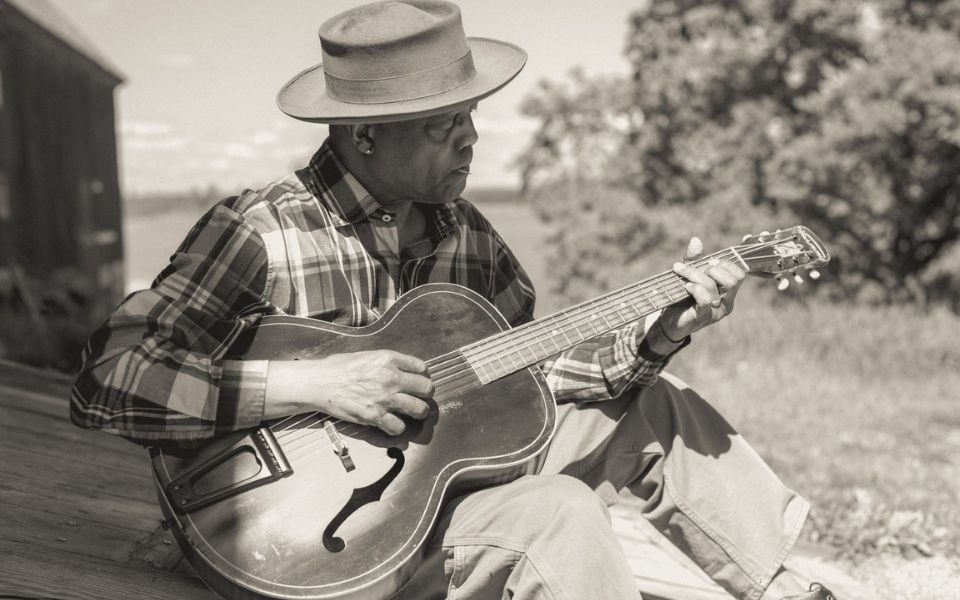 Black and white of Eric Bibb sat outdoors playing a guitar, wearing a plaid shirt and hat shading his face.