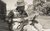 Eric Bibb Black and white of Eric Bibb sat outdoors playing a guitar, wearing a plaid shirt and hat shading his face.