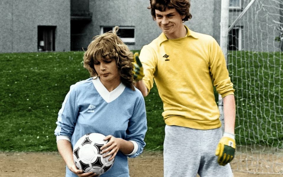A smiling young man in a yellow Umbro shirt and goalie gloves teases a girl holding a football in front of him. They are on the grounds of a housing estate in front of goal posts.