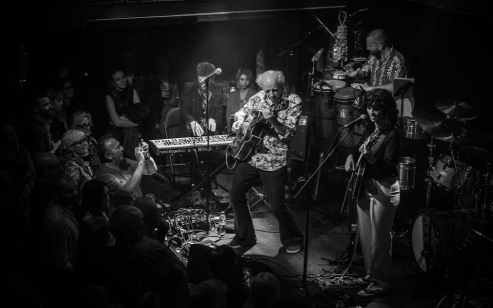 The band Hejira perform in a crowded stage setting. From left to right, there is a keyboard player, guitarist, drummer and guitarist-vocalist beneath the moody spotlight.