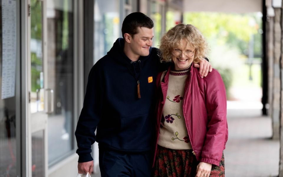 A younger man in a hoodie smiles with an older woman in glasses and a floral knitted jumper who laughs as they walk along a street.