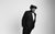 John Kearns Black and white photo of John Kearns in a simple suit, wearing a comic wig and fake teeth. He tilts sideways against a blank backdrop.