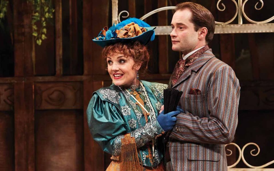 Kerry Ellis dressed in a blue Victorian-style dress and hat, holding hands with Peter Sandys-Clarke dressed in a Victorian striped grey suit in The Importance of Being Earnest.