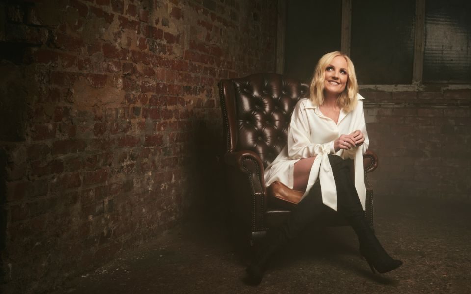 Kerry Ellis sitting in a brown armchair and wearing a long white shirt dress and black over-the-knee boats.