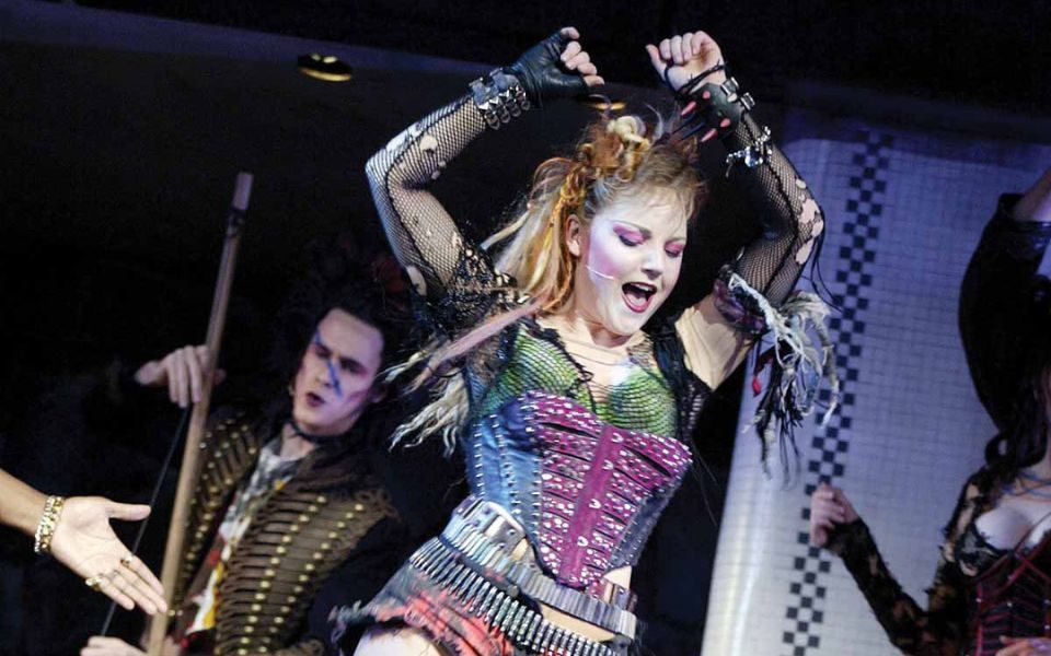 Kerry Ellis dressed at Meat in We Will Rock You. She wears a punky costume with red and black short shorts, belts, a purple and green corset, and fishnet sleeves.