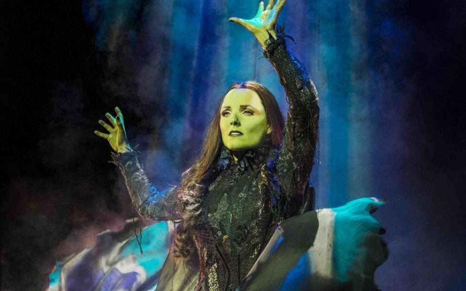 Kerry Ellis as Elphaba in Wicked. She is painted green and wears a long black wig with a black witchy dress.