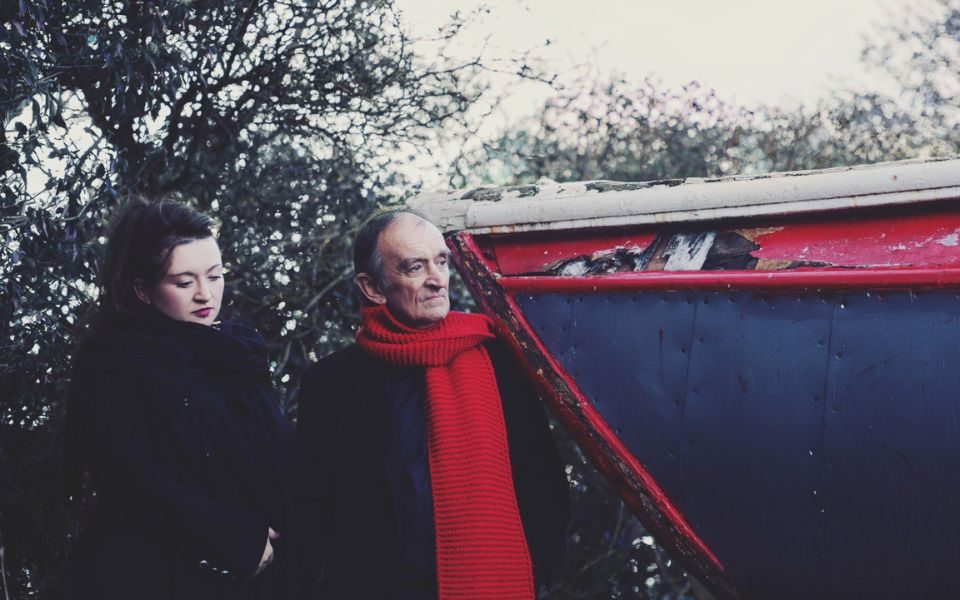 Martin and Eliza Carthy stand together in thick winter coats and long scarfs, both looking into the distance. The background is an icy scene with wintry trees and a dilapidated boat.