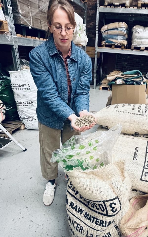 Hols Kragiopoulos stands in a warehouse and scoops coffee beans out of a bag and shows them to camera. She wears a denim jacket and tan trousers.