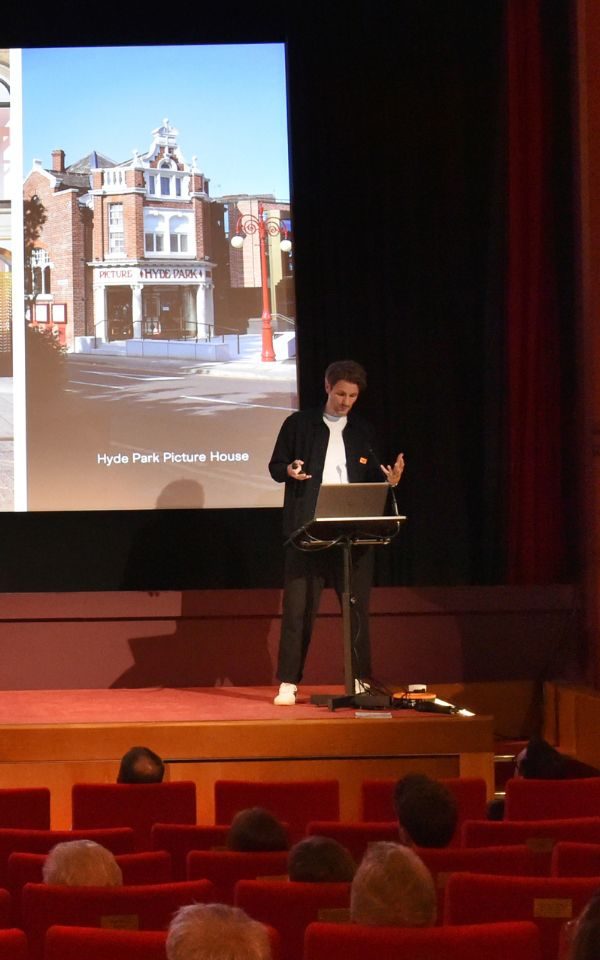 Mark Johnston stands on stage at Hyde Park Picture House delivering a talk. There is an image of the exterior of the venue on the screen behind him.