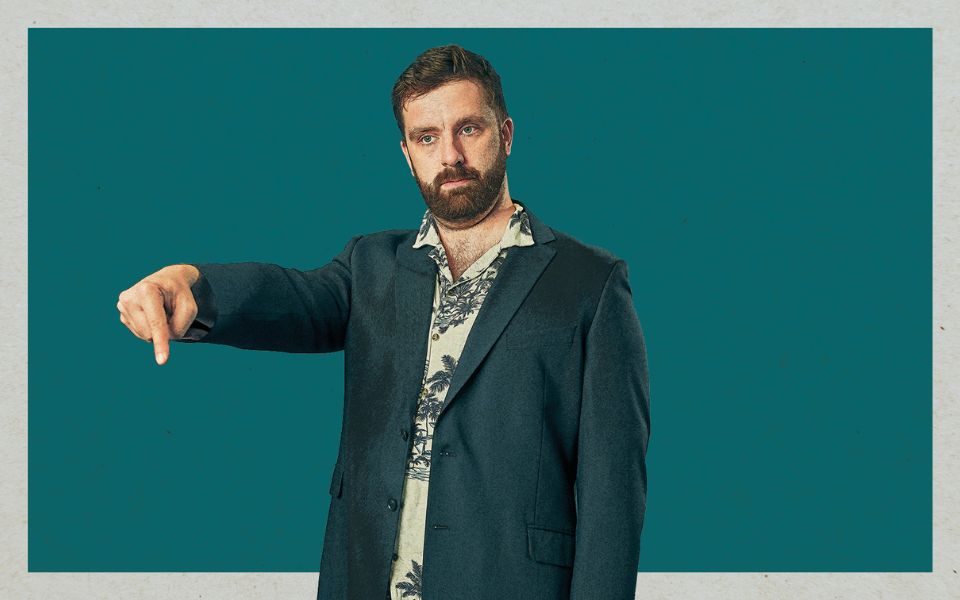 In a forest green suit and shirt patterned with palm trees, a straight-faced Pierre Novellie looks off-camera and points his finger downwards, set against a plain blue-green backdrop.