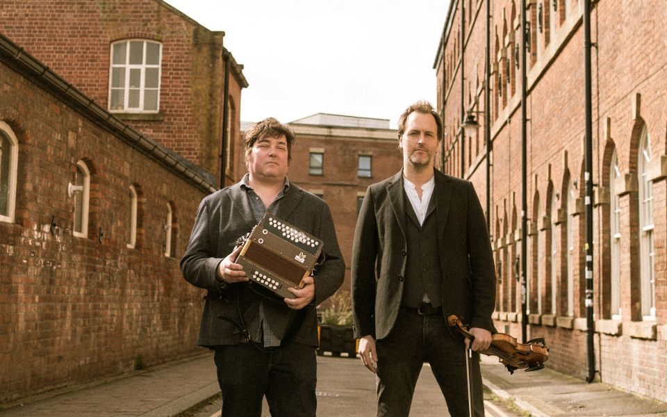 Spiers and Boden stand in the middle of a street of red brick buildings. Both wear casual suit jackets, one holds an accordion and the other a violin.
