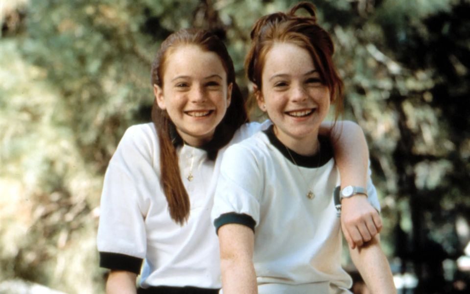Two twins, both Lindsay Lohan, sat smiling together outdoors with their arms round each other.
