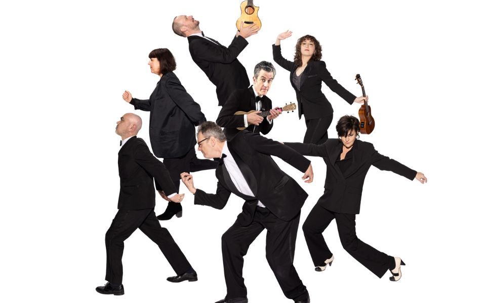 Seven people in matching black suits strut around a plain white backdrop with exaggerated movements, some holding a ukulele .