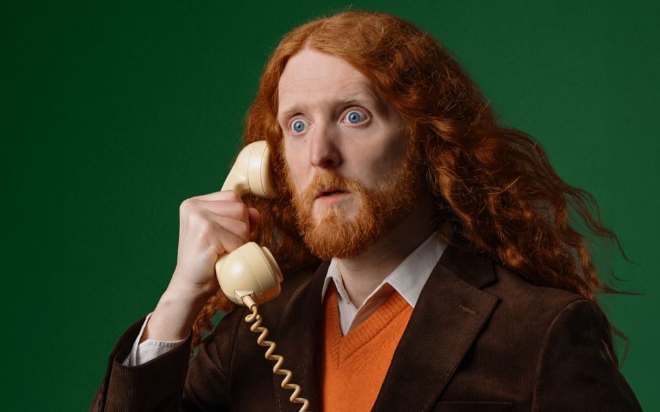 Alasdair Beckett-King's long red hair breezes over his shoulder as he holds a white telephone to his ear with a shocked expression. He wears an orange jumper and brown suit jacket, as he stands in front of a dark green background.