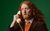 Alasdair Beckett-King Alasdair Beckett-King's long red hair breezes over his shoulder as he holds a white telephone to his ear with a shocked expression. He wears an orange jumper and brown suit jacket, as he stands in front of a dark green background.