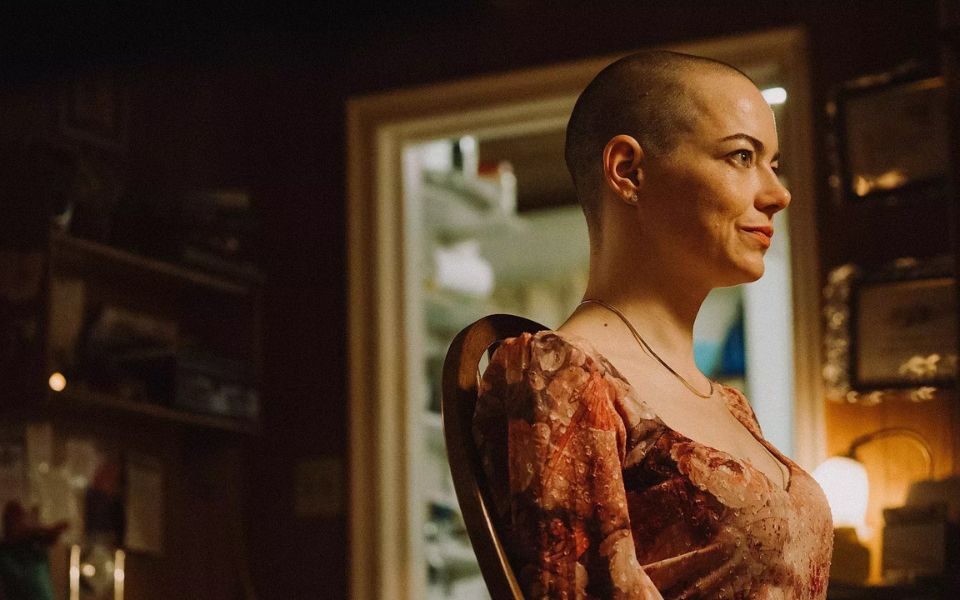 Emma Stone with a shaved head sits in a dress at a wooden chair and looks straight ahead.