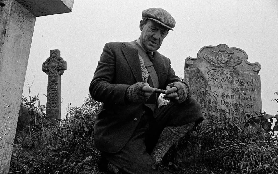 Black and white film still. A man kneels down in an overgrown graveyard, closely examining something in his hands. He wears a flat cap, satchel, tweed suit and fingerless gloves.