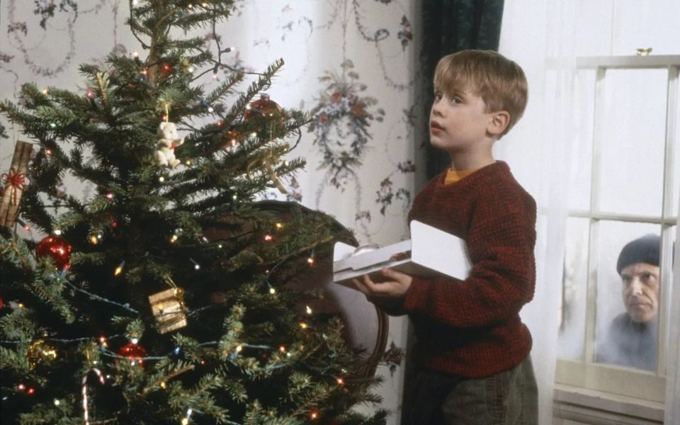 A young boy in a red jumper (Macaulay Culkin) decorates a Christmas tree in a bright living room. Behind him, a man in a black beanie (Joe Pesci) peers through a foggy window.